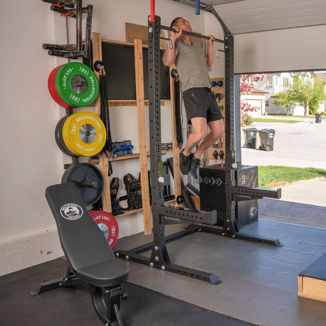 A man uses the Bells of Steel USA Home Gym Builder to do pull-ups in a garage gym with barbells, a weight plate set, and an adjustable bench. Sunlight from the open door reveals houses outside.