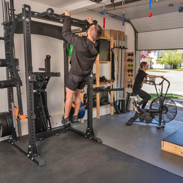 A man does pull-ups on the Bells of Steel USA Home Gym Builder power rack while a woman uses an exercise bike in a well-equipped garage gym with organized weights and neatly stacked plates.