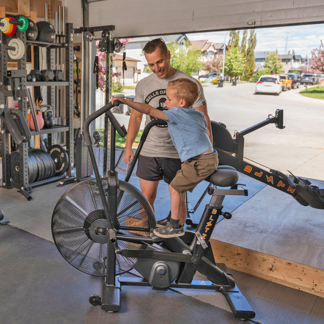 A man smiles next to a boy riding an exercise bike in a garage gym, featuring the Bells of Steel USA Home Gym Builder, power racks, and weight plates, with an open garage door showing a suburban street.
