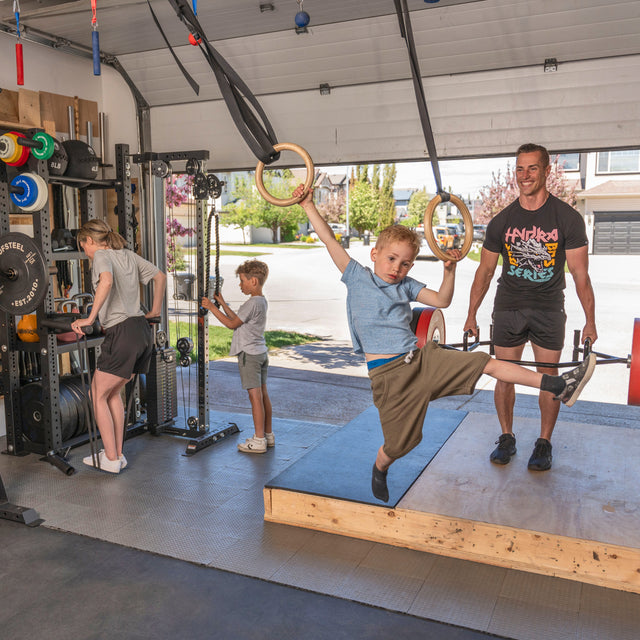 A family enjoys their Bells of Steel USA Home Gym Builder: one boy swings on gymnastic rings, another uses an adjustable bench, a woman lifts weights, and a man supervises as sunlight shines through the open garage door.