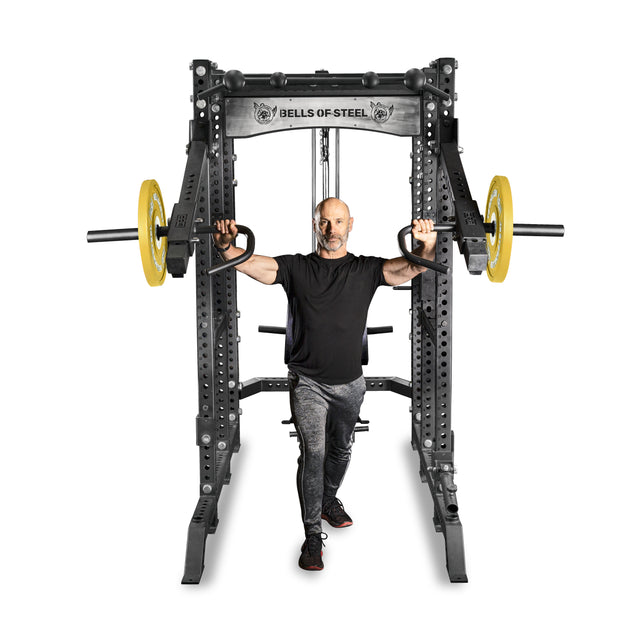 A man in a black t-shirt and gray pants uses the Bells of Steel Lever Arms Rack Attachment, performing a split squat with yellow Olympic weight plates on a white background.