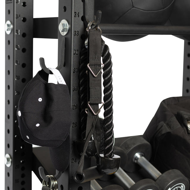 A Bells of Steel Utility Hook holds a black baseball cap, tricep rope, and gym gear on a black metal rack, offering efficient storage with dumbbells and a medicine ball visible in the background.