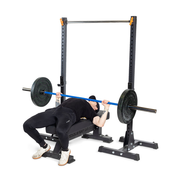 Dressed in a black shirt, black pants, white shoes, and a cap, a person performs bench presses on a bench using the Bells of Steel Multi-Purpose Olympic Barbell – The Utility Bar. A squat rack with safety bars provides maximum utility and performance.