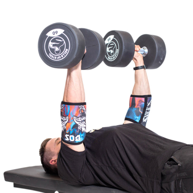 Wearing Bells of Steel Elbow Sleeves with graphic designs, a person presses large dumbbells above their chest while lying on a bench, set against a plain white background.