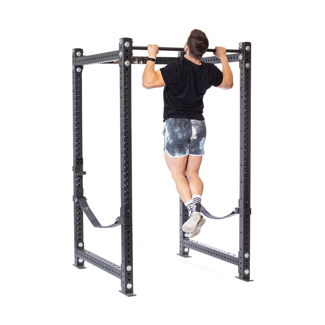 A person does a pull-up on the Bells of Steel Hydra Four Post Power Rack - Prebuilt (3" x 3", ⅝" Holes) with safety straps, making it perfect for any home gym, all set against a white background.
