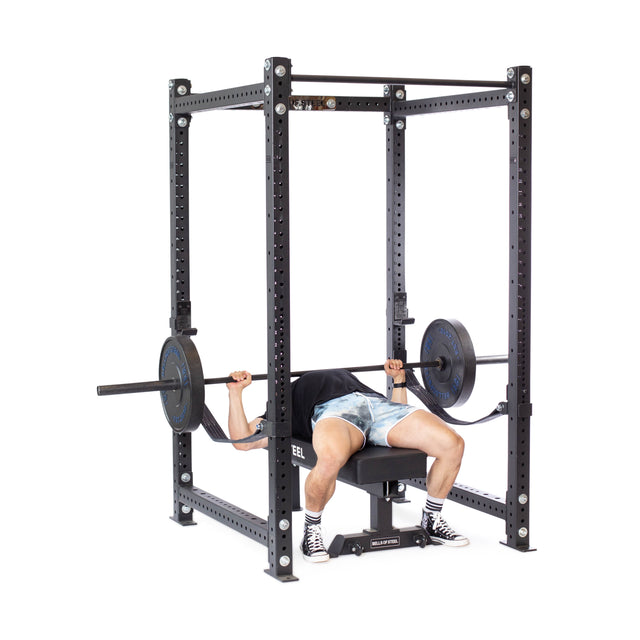 A person bench presses a barbell with weights inside the Bells of Steel Hydra Four Post Power Rack - Prebuilt (3" x 3", ⅝" Holes) in their home gym, wearing shorts and sneakers and gripping the bar above their chest.