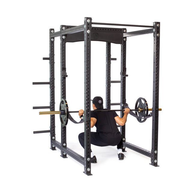 Wearing black workout clothes, a person squats with a barbell loaded with weight plates inside the Bells of Steel Hydra Six Post Power Rack - Prebuilt (3" x 3", ⅝" Holes), which features built-in plate storage pegs.