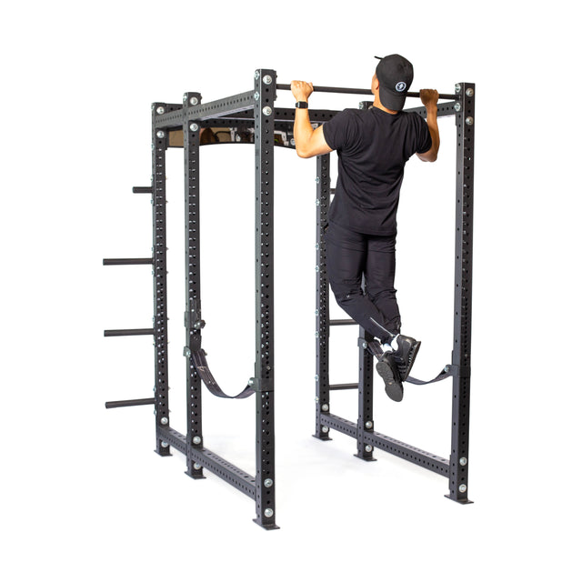 A person in black athletic wear and a cap performs a pull-up on the Bells of Steel Hydra Six Post Power Rack - Prebuilt (3" x 3", ⅝" Holes), featuring multiple attachments and plate storage, set against a white background.