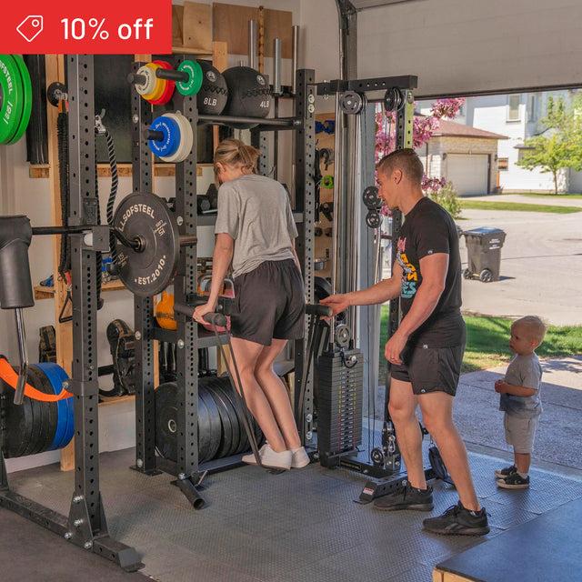 A woman does tricep dips on parallel bars in a garage gym while a man supervises and a child watches. Weightlifting gear is neatly organized on the Bells of Steel Hydra Storage Builder. A "10% off" banner appears in the top left corner.