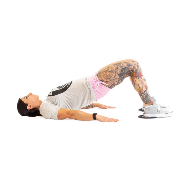 Wearing a gray shirt, pink shorts, and gray shoes, someone with leg tattoos uses Bells of Steel Core Sliders for a total body workout, performing glute bridges on their back with feet on the sliders and hips lifted.
