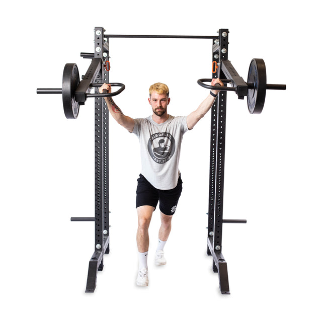 A man with blond hair, in a gray t-shirt and black shorts, performs a lunge holding the handles of Bells of Steel Lever Arms Rack Attachment loaded with weights, attached to a squat rack. The background is plain white.