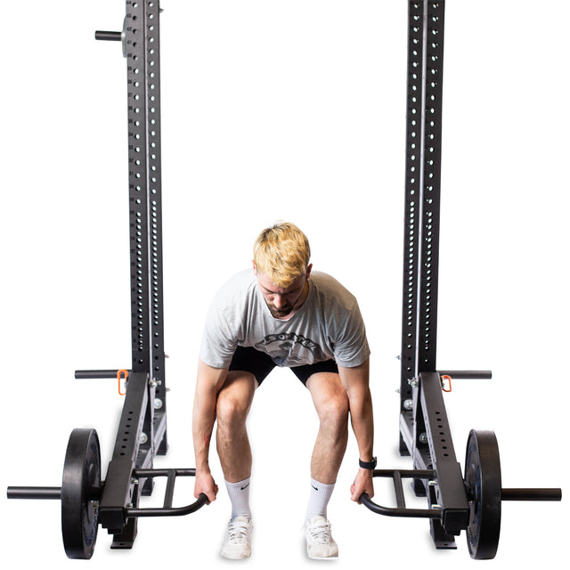 A person with blond hair in a gray t-shirt, black shorts, and white shoes is preparing to use the Bells of Steel Lever Arms Rack Attachment inside a power rack, standing ready in the deadlift position.