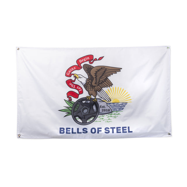 The Bells of Steel Flags feature a white design with an eagle holding a barbell plate, beneath a red "Let Gains Begin" banner. The eagle stands by water and grass, sunrise behind, and the flag reads "BELLS OF STEEL"—ideal gym decor.