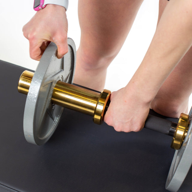 A person attaches a large silver weight plate to the Bells of Steel Loadable Dumbbell Handle while standing beside a black workout bench. Only their hands and lower legs are visible—ideal for any home gym.