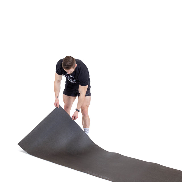 A person is unrolling a large black Bells of Steel Cardio Machine Floor Mat on the floor, wearing a black shirt and shorts against a white background.