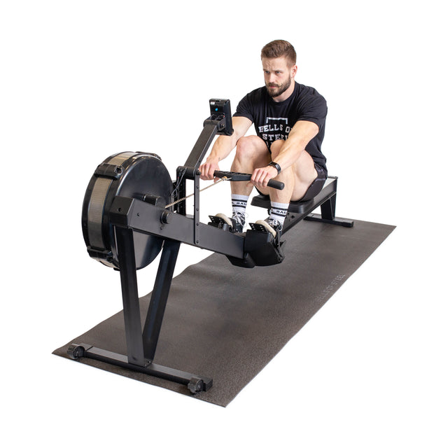 A man in athletic wear uses a rowing machine on a Bells of Steel Cardio Machine Floor Mat, gripping the handle and getting ready to row against a plain white background.