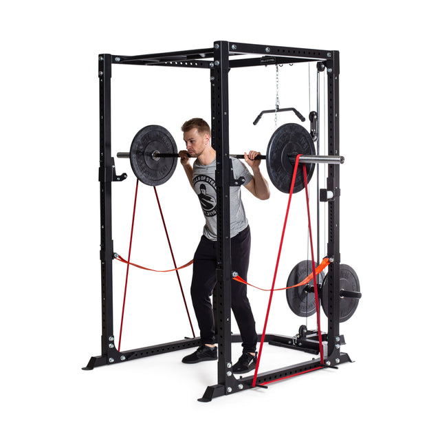 A man does a barbell squat in a black power rack using Bells of Steel Fabric Non-Slip Resistance Bands (41"). Weight plates are loaded on the bar and storage pegs, showcasing powerlifting techniques on a plain white background.
