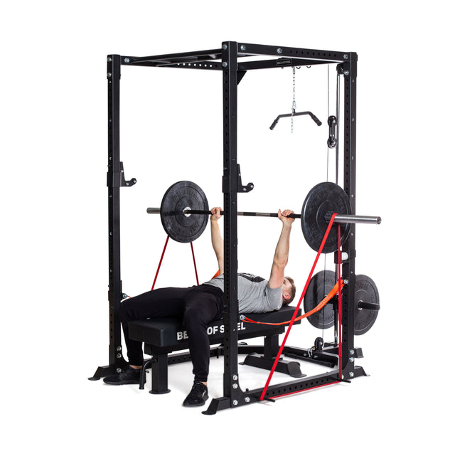 A person in a gray shirt and black pants does a barbell bench press in a power rack, using Bells of Steel 41" Fabric Non-Slip Resistance Bands attached to the bar and rack for added tension—perfect for powerlifting and mobility work.