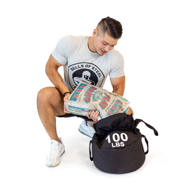 A man in athletic wear kneels and pours sand into a black Bells of Steel Fitness Sandbag Set labeled "100 LBS," highlighting its durable construction against a white background.