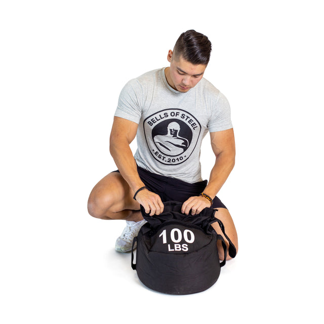A man in a gray Bells of Steel t-shirt kneels on one knee, handling a large black 100 LBS Fitness Sandbag Set by Bells of Steel, designed for sandbag workouts with durable Condura construction, shown against a white background.