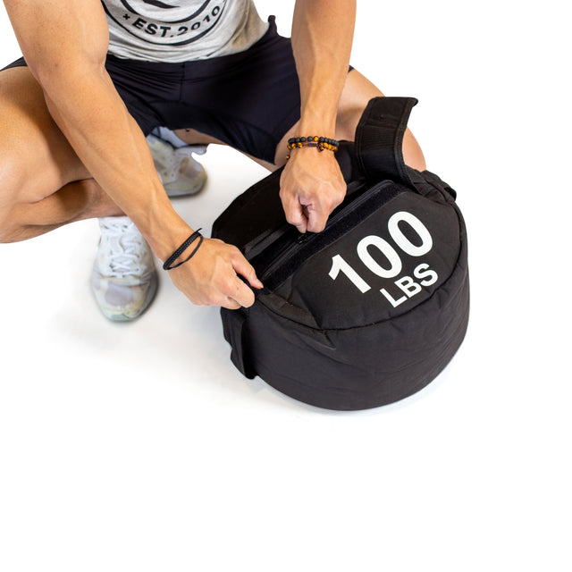 Wearing athletic gear, a person crouches to adjust the strap on a black Bells of Steel Fitness Sandbag labeled "100 LBS," made for intense workouts, set against a white background.