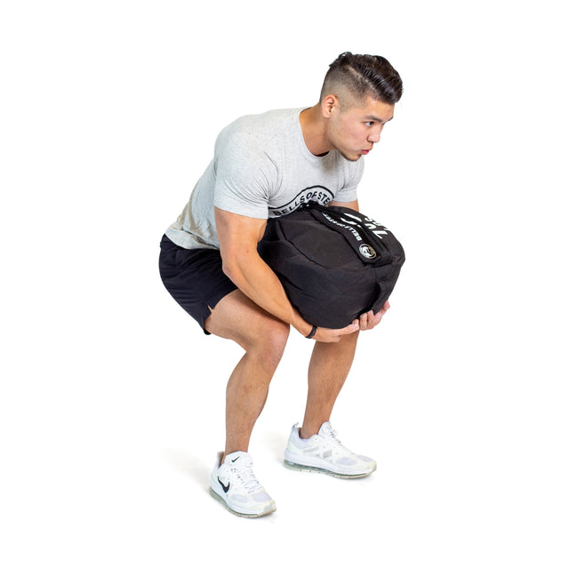 A man, dressed in a gray t-shirt, black shorts, and white sneakers, squats while lifting Bells of Steel Fitness Sandbag Sets with both hands, focusing on his workout against a plain white background.