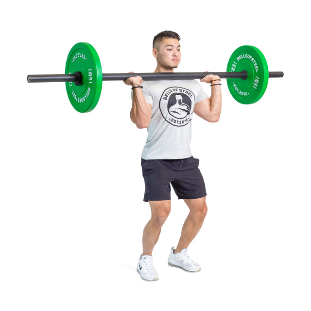A man in a gray t-shirt and black shorts holds Bells of Steel Axle Bars with green plates at shoulder height, ready to lift. This strongman training exercise is ideal for building grip strength. Background is plain white.