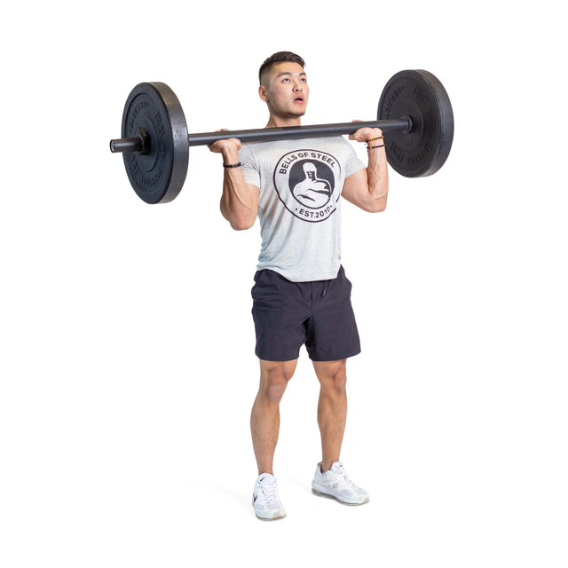 A man in a gray Bells of Steel tee, black shorts, and white sneakers stands with Axle Bars loaded on his shoulders, ready to lift—ideal for grip strength and strongman training. White background.