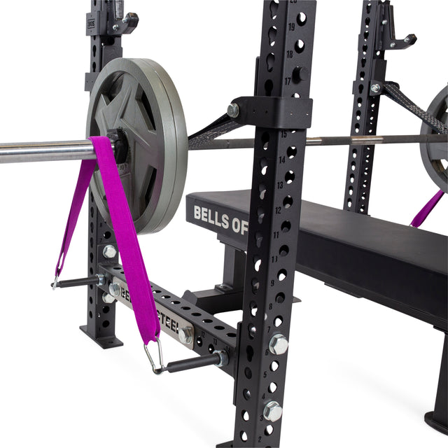 A weightlifting bench press setup features a barbell with weight plates, secured by bright purple safety straps on a black power rack equipped with Bells of Steel Band Pegs with Carabiners for adding resistance bands.