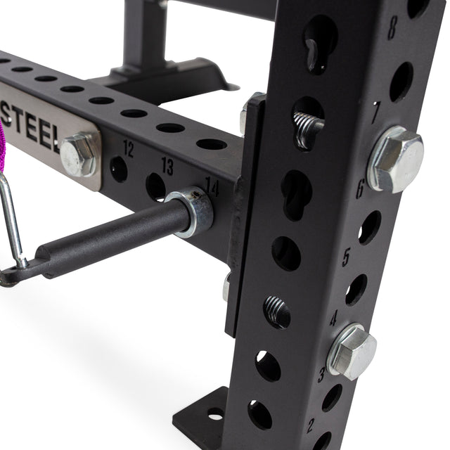 Close-up of Bells of Steel Band Pegs with Carabiners, attached to a black metal power rack with numbered holes and bolts. A "STEEL" label is visible on the frame, along with bar-secured band pegs for resistance bands.