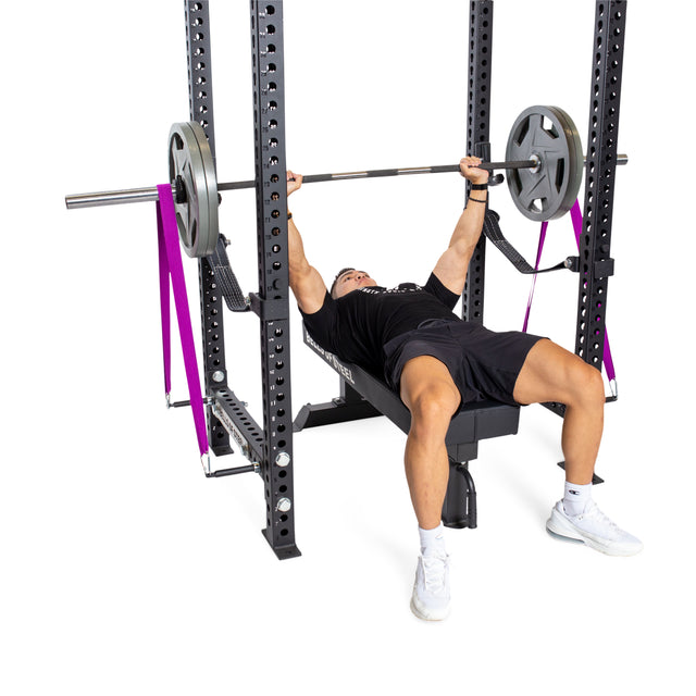 A person in athletic clothing and white sneakers bench presses a barbell inside a power rack. Bells of Steel Band Pegs with Carabiners secure purple resistance bands to the rack for added resistance.