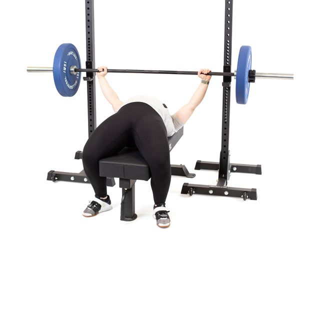 Wearing black leggings and a light shirt, a person bench presses on a bench using a barbell with blue plates, supported by Bells of Steel Squat Stands (2.3" x 2.3", ⅝" holes)—ideal for any home gym setup.