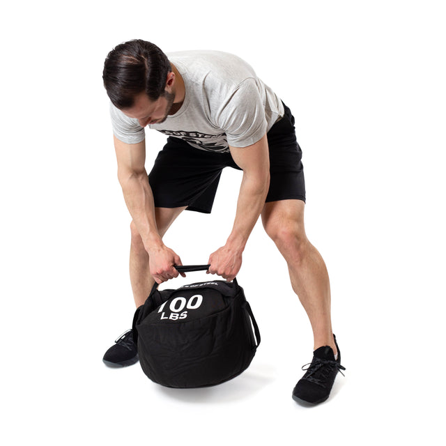 A man in a gray t-shirt and black shorts bends over to grip the handle of a black Bells of Steel Fitness Sandbag Sets labeled "100 LBS," getting ready for his sandbag workout.