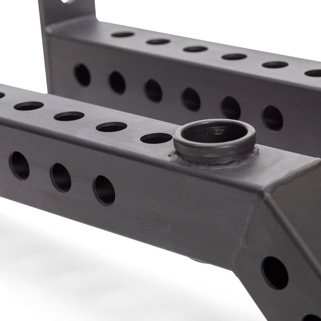 Close-up view of Bells of Steel Front Foot Stabilizers, a black metal power rack frame with rows of circular holes and a cylindrical attachment, designed for enhanced stability during gym or exercise routines.