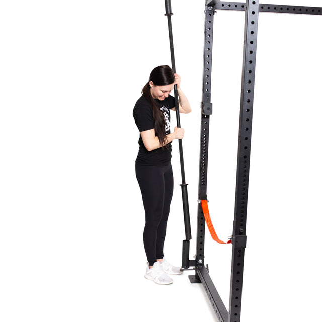 A woman in athletic wear stands by a black squat rack, holding a barbell stored with the Bells of Steel Vertical Mount Barbell Holder Rack Attachment. The minimal white background highlights efficient storage.