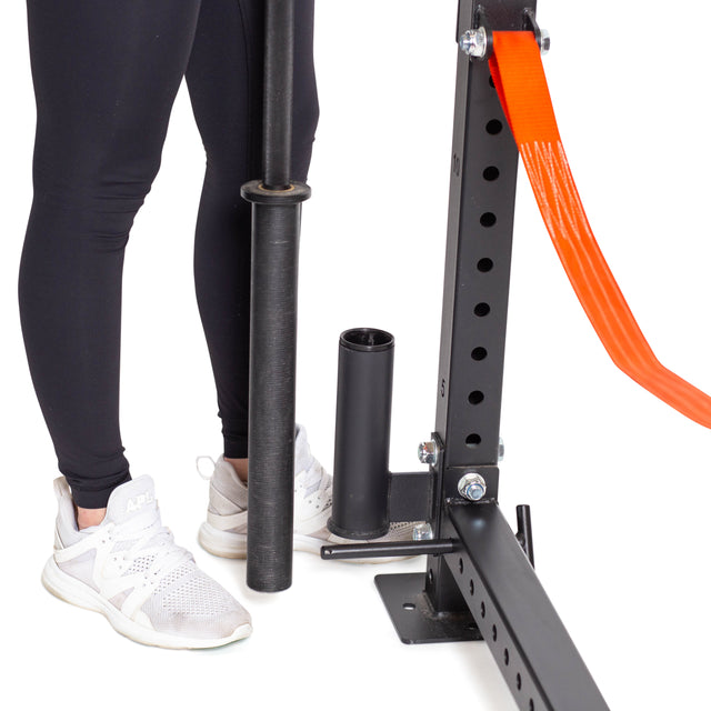 A person in black leggings and white sneakers stands by fitness equipment, holding a barbell near a squat rack with an orange strap, featuring the Bells of Steel Vertical Mount Barbell Holder Rack Attachment for organized storage.