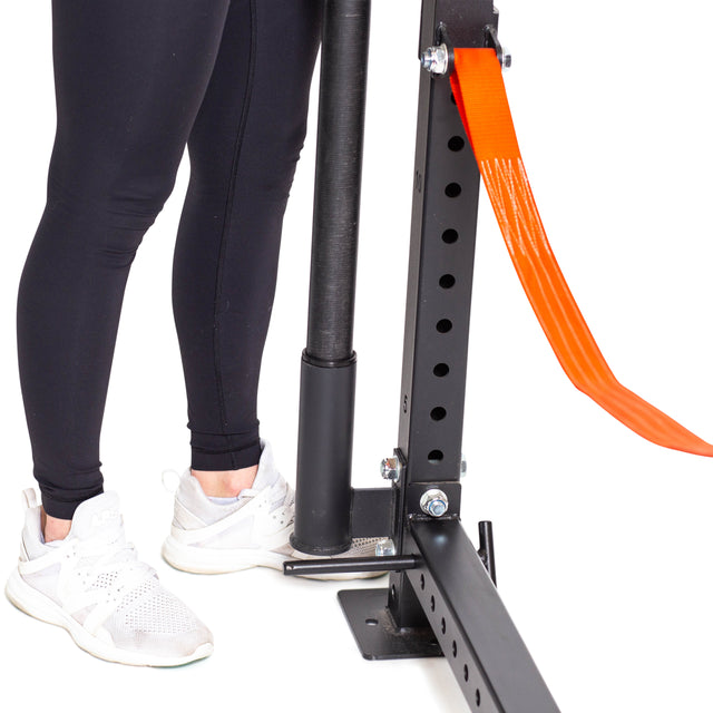 A person in black leggings and white sneakers stands by a Bells of Steel power rack equipped with a Vertical Mount Barbell Holder Rack Attachment and orange safety strap for organized barbell storage.