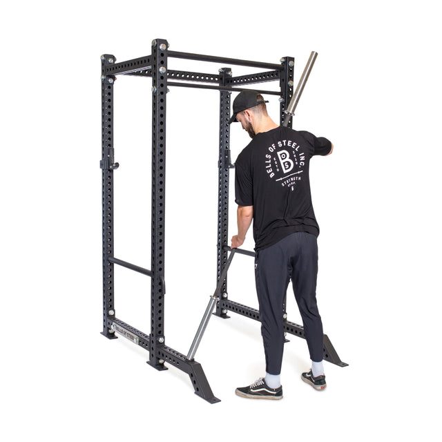 A man in a black "Bells of Steel" shirt attaches Front Foot Stabilizers by Bells of Steel to a power rack, preparing for his workout.