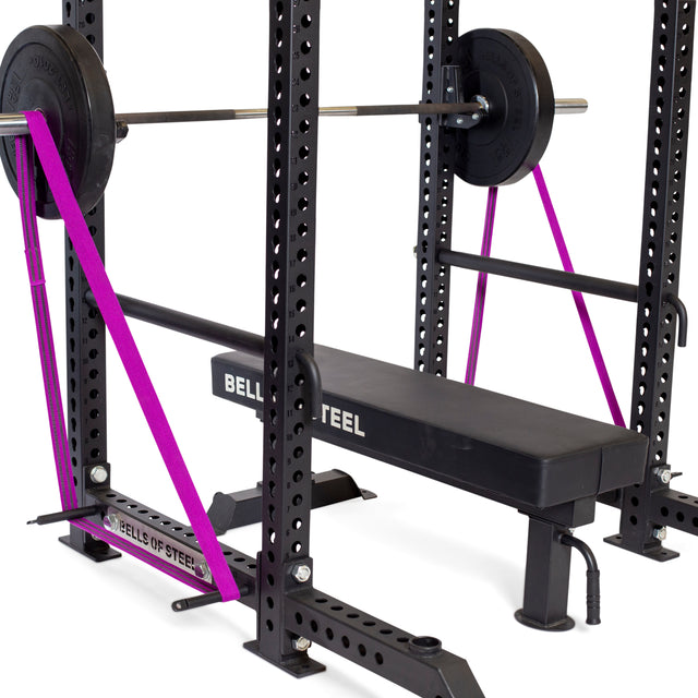 A weight bench sits inside a Bells of Steel power rack with a barbell and weights. Two purple resistance bands are looped on Standard Band Pegs attached to the rack’s base and barbell. All equipment is from Bells of Steel.