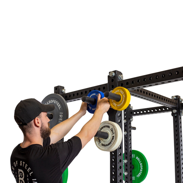 A person in a black cap and t-shirt adjusts a yellow weight plate on the power rack, which is equipped with Bells of Steel's Change Plate Storage Pegs, featuring Olympic change plates in blue and white against a plain white background.