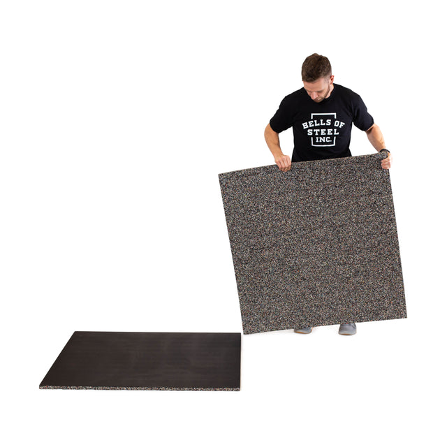 A person in a Bells of Steel t-shirt lifts a large, rectangular Rubber Flooring Gym Mat 39" x 39", with another mat from the same brand lying flat nearby.