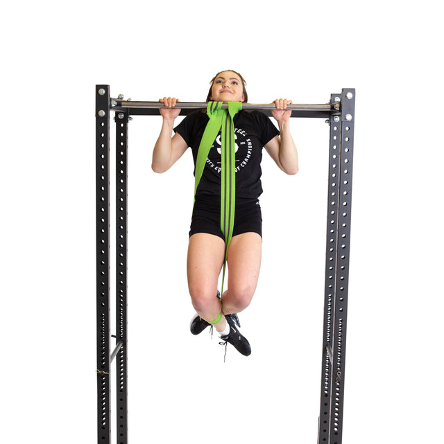 A woman uses the Bells of Steel Fabric Non-Slip Resistance Band (41”) looped around a metal bar and her legs to assist with pull-ups. She wears black workout attire against a white background—ideal for mobility or powerlifting routines.