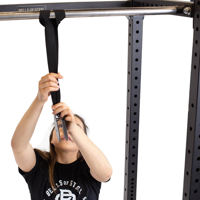 A person wearing a black shirt adjusts the strap on a Bells of Steel cable pulley attached to a metal pull-up bar within the gym's power rack, all against a plain white background that emphasizes precision and strength.