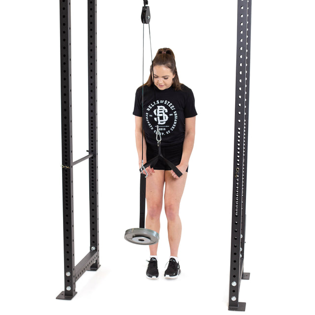 A woman is using a Bells of Steel Cable Pulley on a power rack for a tricep pushdown. She's in profile, wearing a black T-shirt and shorts, focused within the metal frame structure.
