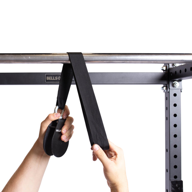A person fastens the Bells of Steel Cable Pulley, which has a black strap and round handle, to a metal pull-up bar on a workout rack to set up portable tricep pushdowns and other exercises.