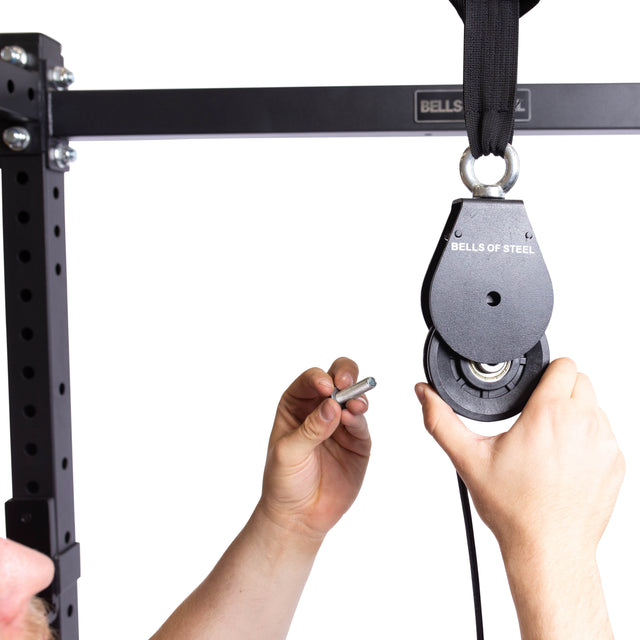 Close-up of hands installing a Bells of Steel Cable Pulley onto a black metal rack with a metal pin against a white background—this portable setup is perfect for tricep pushdowns and other gym exercises.