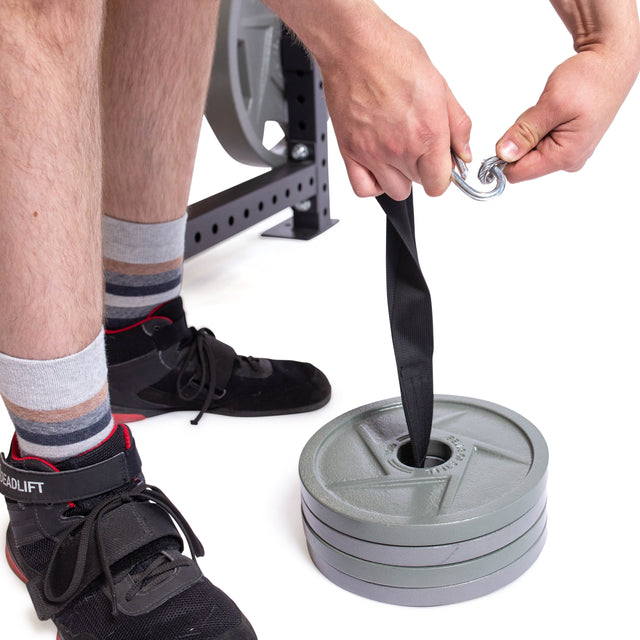 Wearing black and red shoes with striped socks, a person tests the weight plate capacity by attaching a carabiner to the strap of a Bells of Steel Cable Pulley. They sit beside gym equipment, preparing for a weightlifting exercise.