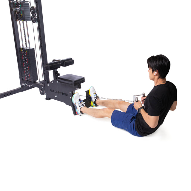 A person in a black shirt and blue shorts performs seated cable rows using the Bells of Steel Double D Handle Cable Attachment, gripping both handles with feet braced on the platform.