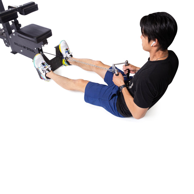 A person in a black shirt and blue shorts sits on the floor using a rowing machine, pulling the Bells of Steel Double D Handle Cable Attachment with both hands while their feet are secured on the footrests.