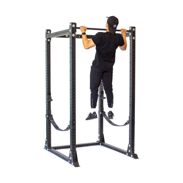 A person in black workout gear performs a pull-up using resistance straps on the Bells of Steel Hydra Flat Foot Power Rack - Prebuilt (3" x 3", ⅝" Holes), perfect for home gyms, isolated on a white background.